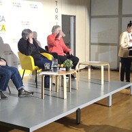 Kobieta przy mównicy przemawia do publiczności, a trzej mężczyźni na scenie słuchają. Jeden z nich pije wodę.