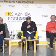 Trzech mężczyzn siedzi na scenie na tle ścianki z logo „Książnica Podlaska”. Środkowy czyta na głos, trzymając mikrofon i książkę. Po bokach stoliki z wodą i roślinami.