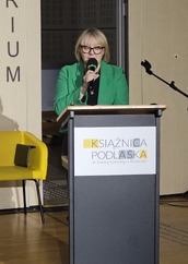 Kobieta w zielonej marynarce mówi do mikrofonu, stojąc przy mównicy z logo „Książnica Podlaska”. W tle żółte krzesło.