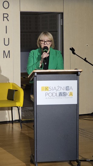 Kobieta w zielonej marynarce mówi do mikrofonu, stojąc przy mównicy z logo „Książnica Podlaska”. W tle żółte krzesło.