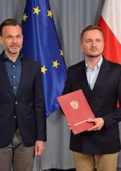 Dwaj mężczyźni w garniturach stoją na tle flag Polski i Unii Europejskiej; jeden z nich trzyma czerwoną teczkę z godłem i napisem „Wojewoda Podlaski”.