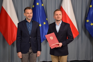 Dwaj mężczyźni w garniturach stoją na tle flag Polski i Unii Europejskiej; jeden z nich trzyma czerwoną teczkę z godłem i napisem „Wojewoda Podlaski”.