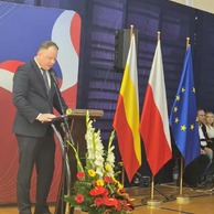 Pionowe zdjęcie przedstawiające mężczyznę w garniturze, Marcin Piętka, przemawiającego z podium, z widocznymi flagami Białegostoku, Polski i Unii Europejskiej.