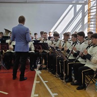 Zdjęcie przedstawiające młodzieżową orkiestrę dętą grającą na instrumentach, prowadzona przez dyrygenta stojącego tyłem do widza.