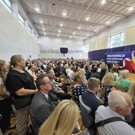 Zdjęcie przedstawiające publiczność, która siedzi na krzesłach w dużej sali, zwrócona w stronę sceny z transparentem