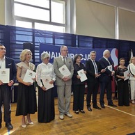 Zdjęcie przedstawia grupę dziewięciu dorosłych osób stojących w rzędzie na scenie. Wszyscy trzymają w rękach białe dokumenty, prawdopodobnie dyplomy lub wyróżnienia. W tle widać niebieską ściankę ze wzorem.