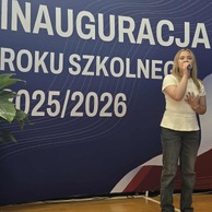 Zdjęcie przedstawiające młodą kobietę śpiewającą do mikrofonu na scenie, z transparentem z napisem