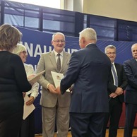 Zdjęcie przedstawiające grupę ludzi na scenie, gdzie jeden z mężczyzn w garniturze przyjmuje dokument, prawdopodobnie dyplom lub wyróżnienie.