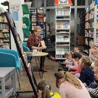 Kobieta w okularach i kratkowanej marynarce siedzi przy małym stoliku, trzymając książkę i uśmiechając się do grupy dziewczynek siedzących na podłodze przed nią w bibliotece.