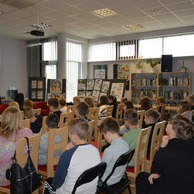 Szeroki plan spotkania autorskiego w bibliotece. Na pierwszym planie widać tyły głów dorosłych i dzieci siedzących na drewnianych krzesłach. Z boku widać kobietę, prawdopodobnie autorkę, stojącą przy projektorze, na którym wyświetla się napis 