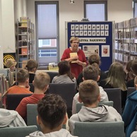 Kobieta w czerwonym swetrze stoi i trzyma książkę, rozmawiając z grupą młodzieży i dzieci siedzących na krzesłach w bibliotece. Za nią widoczny jest plakat z napisem 