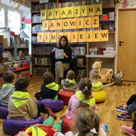 Kobieta czyta książkę grupie małych dzieci siedzących na poduszkach na podłodze w bibliotece. Za kobietą na półce widać duże żółte litery tworzące imiona i nazwisko: Katarzyna Janowicz Timoﬁejew.