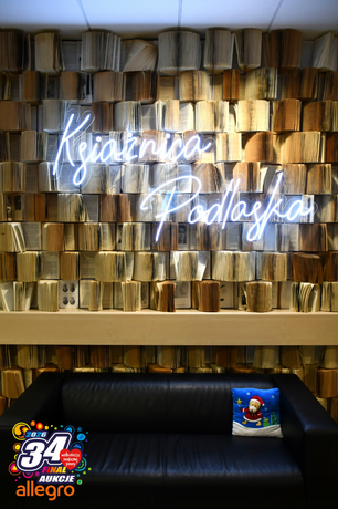 Wnętrze Książnicy Podlaskiej z artystyczną instalacją - ściana wypełniona setkami pionowo ułożonych, otwartych książek tworzących unikatowe tło. Na tym tle świeci niebieski neon z napisem 'Książnica Podlaska'. Przed instalacją stoi czarna skórzana sofa z niebieską poduszką z motywem świątecznym. W lewym dolnym rogu widoczne kolorowe logo 34. Finału Wielkiej Orkiestry Świątecznej Pomocy z napisem 'Aukcje Allegro'."