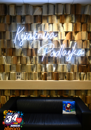 Wnętrze Książnicy Podlaskiej z artystyczną instalacją - ściana wypełniona setkami pionowo ułożonych, otwartych książek tworzących unikatowe tło. Na tym tle świeci niebieski neon z napisem 'Książnica Podlaska'. Przed instalacją stoi czarna skórzana sofa z niebieską poduszką z motywem świątecznym. W lewym dolnym rogu widoczne kolorowe logo 34. Finału Wielkiej Orkiestry Świątecznej Pomocy z napisem 'Aukcje Allegro'."