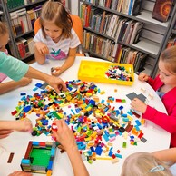 Zdjęcie nr 6 przedstawia dzieci siedzące  przy okrągłym, białym stole. Na stole są rozsypane klocki LEGO a w tle są regały z książkami.