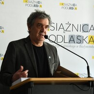 Dariusz Szada-Borzyszkowski z Galerii im. Sleńdzińskich przemawia przy mównicy. W tle baner z logo Książnicy Podlaskiej.