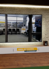 Kobieta stoi w wirtualnym studiu telewizyjnym przed dużym ekranem. Ekran pokazuje wnętrze jakiegoś pomieszczenia, być może poczekalni, z rzędem czarnych krzeseł. Kobieta ma na sobie jasny sweter i szare spodnie. Stoi po prawej stronie ekranu. Pod ekranem znajduje się szafka RTV z głośnikami po bokach, a na niej widoczna jest plakietka z napisem ANNA KOCHANIEC i pod spodem mniejszymi literami DZIAŁ ZESPOŁÓW SPECJALNYCH. Ściana za ekranem wygląda jak ściana z czerwonej cegły. Po obu stronach mebla RTV i ekranu znajdują się doniczkowe rośliny. U góry po prawej stronie widać logo z napisem KSIĄŻNICA PODLASKA. Podłoga jest drewniana.