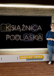 Kobieta stoi w wirtualnym studiu telewizyjnym przed dużym ekranem. Ekran wyświetla logo/napis neonowy KSIĄŻNICA PODLASKA. Kobieta ma na sobie ciemnoniebieską bluzkę i jasne dżinsy. Stoi po prawej stronie ekranu, trzymając złożone ręce przed sobą. Pod ekranem znajduje się szafka RTV z głośnikami po bokach, a na niej widoczna jest plakietka z napisem MARTA KOZŁOWSKA i pod spodem mniejszymi literami WYPOŻYCZALNIA GŁÓWNA I MIĘDZYBIBLIOTECZNA. Ściana za ekranem wygląda jak ściana z czerwonej cegły. Po obu stronach mebla RTV i ekranu znajdują się doniczkowe rośliny. U góry po prawej stronie widać logo z napisem KSIĄŻNICA PODLASKA. Podłoga jest drewniana.