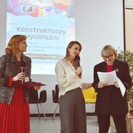 Trzy kobiety na scenie. Po lewej prowadząca w czerwieni, w środku kobieta w jasnej bluzce, po prawej kobieta w okularach uśmiecha się, patrząc w dokumenty.