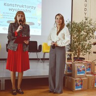 Dwie kobiety na scenie w auli. Po lewej kobieta w czerwonej spódnicy przemawia do mikrofonu, po prawej kobieta w białej bluzce i szarych spodniach. W tle plakat konkursu „Konstruktorzy wyobraźni”.