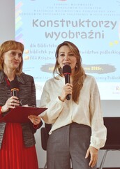 Zbliżenie na dwie kobiety przemawiające do mikrofonów. Po lewej kobieta w czerwonej spódnicy i żakiecie, po prawej w jasnej, koronkowej bluzce.