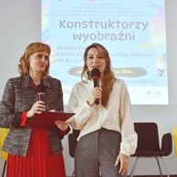 Zbliżenie na dwie kobiety przemawiające do mikrofonów. Po lewej kobieta w czerwonej spódnicy i żakiecie, po prawej w jasnej, koronkowej bluzce.