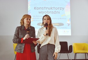 Zbliżenie na dwie kobiety przemawiające do mikrofonów. Po lewej kobieta w czerwonej spódnicy i żakiecie, po prawej w jasnej, koronkowej bluzce.