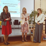 Trzy prelegentki na scenie. Kobieta w środku, w kwiecistej sukience i jasnym żakiecie, czyta z kartki do mikrofonu. Po prawej widoczne podium z logo Książnicy Podlaskiej.
