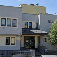 Widok frontowy budynku Filii nr 9 Książnicy Podlaskiej przy ul. Gajowej 73 w Białymstoku, uchwycony od strony ulicy. Dwupiętrowy budynek z jasną elewacją i napisem „BIBLIOTEKA
