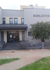 Frontowa elewacja budynku Filii nr 9 Książnicy Podlaskiej przy ul. Gajowej 73 w Białymstoku. Dwupiętrowy budynek z jasnoszarą elewacją, z widocznym napisem „BIBLIOTEKA" w prawej górnej części fasady. Na pierwszym planie wejście główne z szerokimi schodami i podjazdem dla niepełnosprawnych, ogrodzenie z metalowych prętów oraz zaparkowany rower dziecięcy. Po prawej stronie wejścia rośnie rozłożyste drzewo liściaste. Zdjęcie wykonane od strony chodnika, w dzień, przy zachmurzonym niebie.