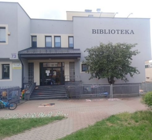 Frontowa elewacja budynku Filii nr 9 Książnicy Podlaskiej przy ul. Gajowej 73 w Białymstoku. Dwupiętrowy budynek z jasnoszarą elewacją, z widocznym napisem „BIBLIOTEKA" w prawej górnej części fasady. Na pierwszym planie wejście główne z szerokimi schodami i podjazdem dla niepełnosprawnych, ogrodzenie z metalowych prętów oraz zaparkowany rower dziecięcy. Po prawej stronie wejścia rośnie rozłożyste drzewo liściaste. Zdjęcie wykonane od strony chodnika, w dzień, przy zachmurzonym niebie.