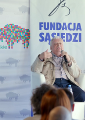 Andrzej Sapkowski siedzi w białym fotelu podczas publicznego wystąpienia. Jest ubrany w jasną, beżową kurtkę rozpiętą nad koszulą w paski oraz ciemne spodnie, a w dłoni trzyma mikrofon, gestykulując drugą ręką w trakcie mówienia. Za jego plecami widoczne są dwa banery promocyjne: jeden z kolorową grafiką żubra i napisem „Podlaskie”, a drugi z dużą, niebieską nazwą „FUNDACJA SĄSIEDZI”. Mężczyzna znajduje się na podwyższeniu, a na pierwszym planie, u dołu kadru, widać rozmazane sylwetki osób z publiczności obserwujących spotkanie.